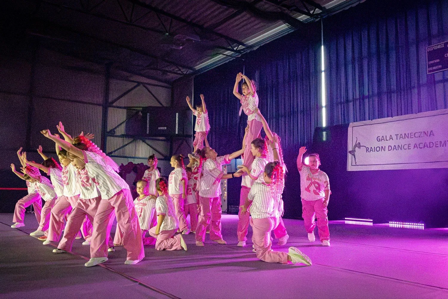 Duża grupa dzieci w strojach scenicznych podczas występu na scenie, w tle baner Gali Tanecznej Raion Dance Academy.